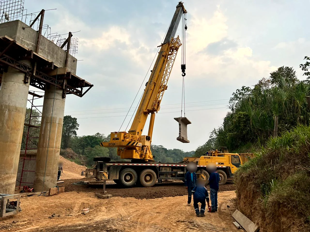 Grúa izando una pieza de hormigón junto a las pilas del puente sobre el río Apere, con personal de obra (rostros difuminados)